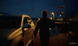 Screenshot of a documentary film showing a woman holding a box and walking past a car.