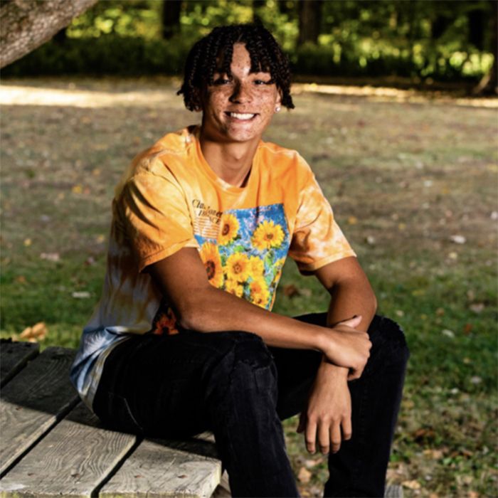 A young man smiles at the camera in a park while sitting on a bench.