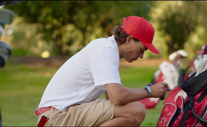 Landon "Happy" Gilmore crouches by his golf bag in a white polo and red hat.