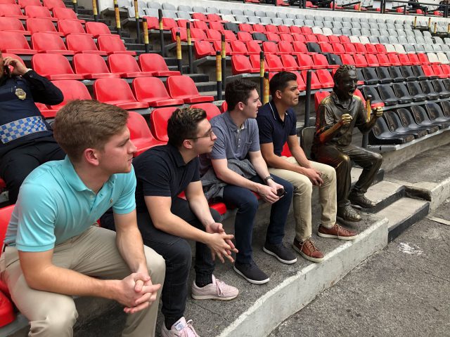 Four men sitting in the front row of bleachers in an empty soccer stadium next to a statue of a screaming fan.