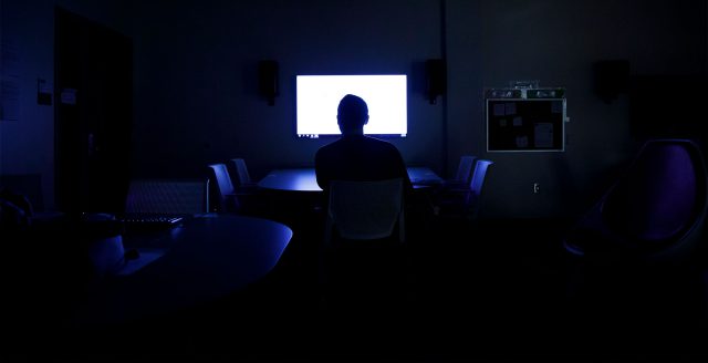 A man sitting in the middle of a room with the lights off looking at a TV screen.