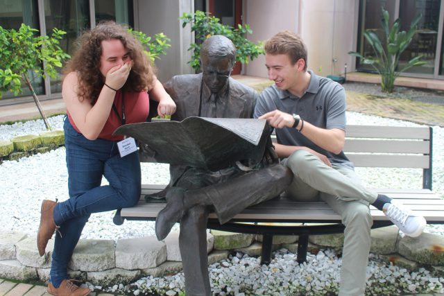 Annie Aguiar and Stefan Krajisnik sit with a statue of a man reading the newspaper sitting on a bench.