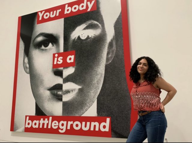 Marille Velez poses next to a mural of a face with the words, "Your body is a battleground."
