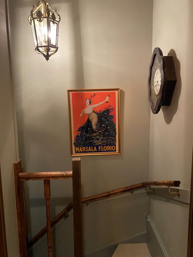 A picture hangs on the wall with a lamp hanging above in a hallway leading to a stairwell.